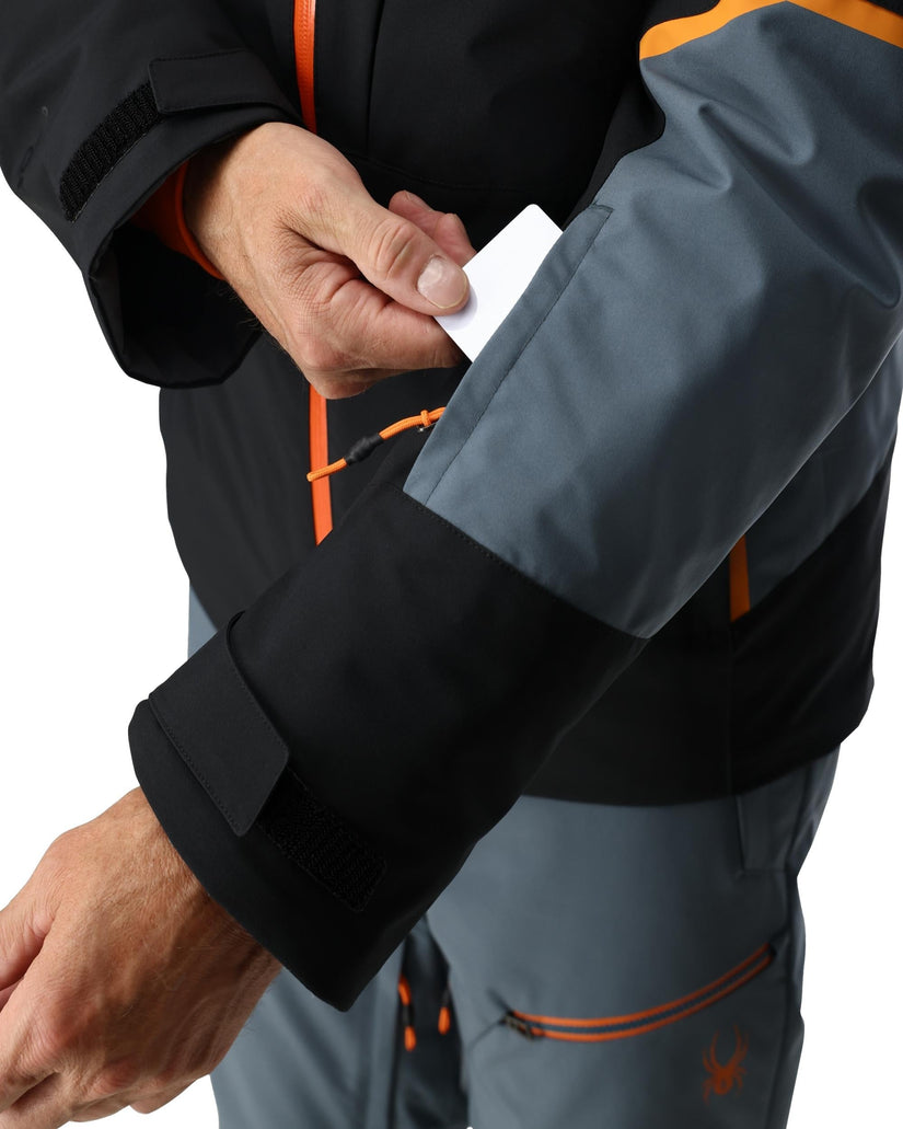 Person wearing a gray and black jacket with orange accents, holding a white card.